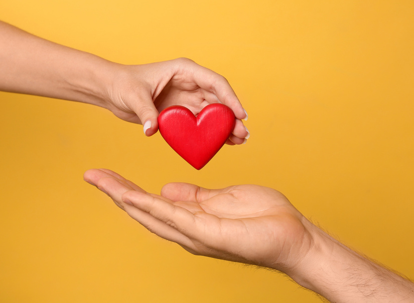 Woman giving red heart to man on yellow background, closeup. Donation concept